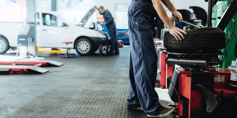 Mechanic replacing tire of while on machine and other inspecting a vehicle.